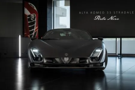 Front view of a gray Alfa Romeo 33 Stradale in a showroom, headlights on, 'Perla Nera' banner in the background