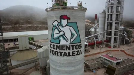 Industrial cement plant with a tall silo displaying the Cementos Fortaleza logo (worker holding a hammer) against a rugged landscape.
