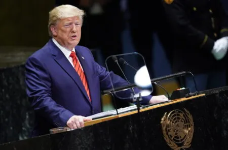 A man in a blue suit and red tie speaks at a United Nations podium with a clear protective screen and microphones in front of him.