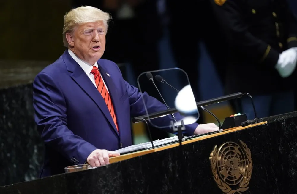 A man in a blue suit and red tie speaks at a United Nations podium with a clear protective screen and microphones in front of him.