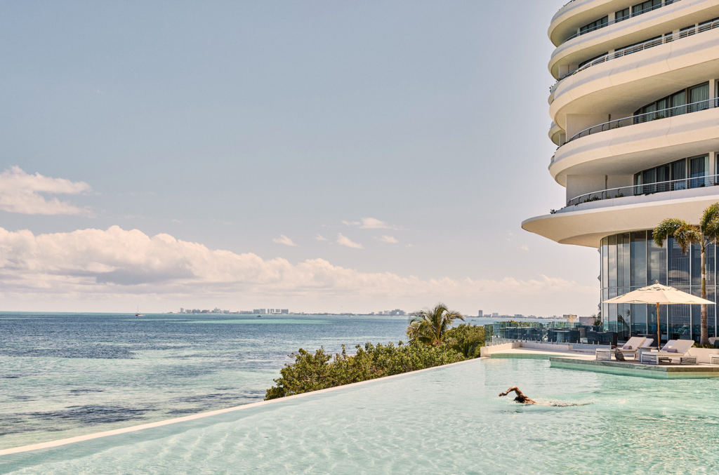 SHA_2 - Petróleo y Energía White modern hotel with curved, stacked balconies on the right, overlooking an infinity pool and calm blue ocean under a clear sky.