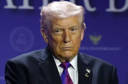 Serious-looking man with light hair in a dark suit and purple tie, seated on a stage with a blue backdrop.