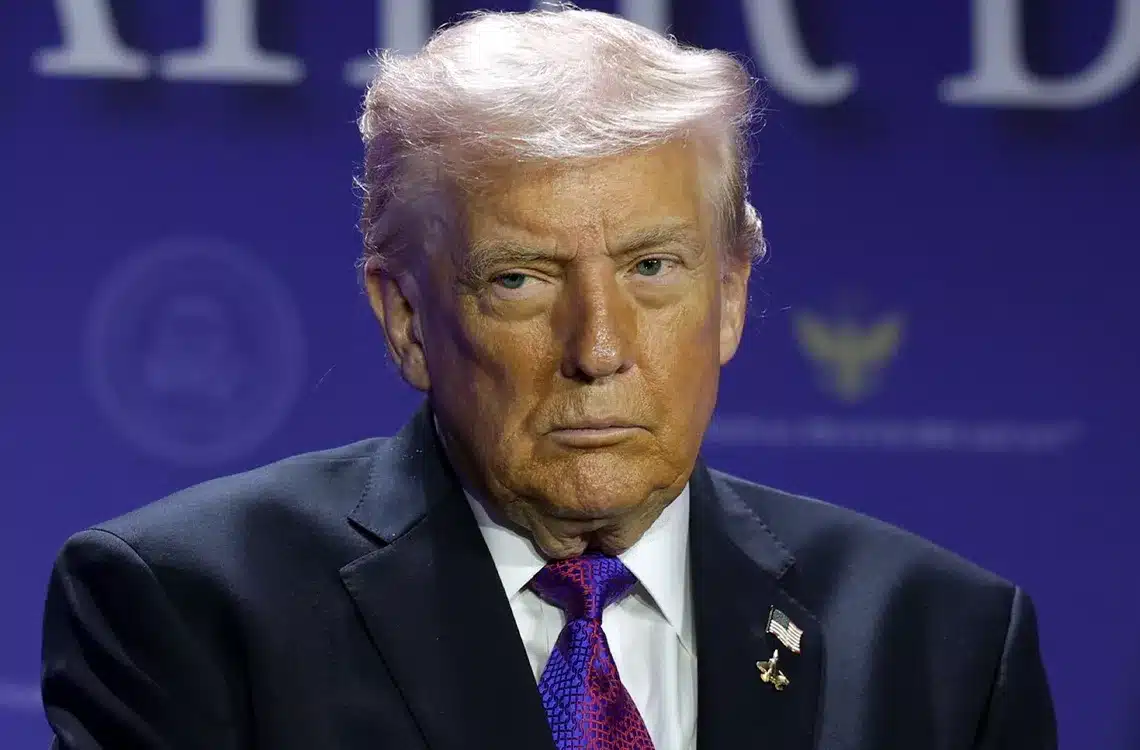 Serious-looking man with light hair in a dark suit and purple tie, seated on a stage with a blue backdrop.