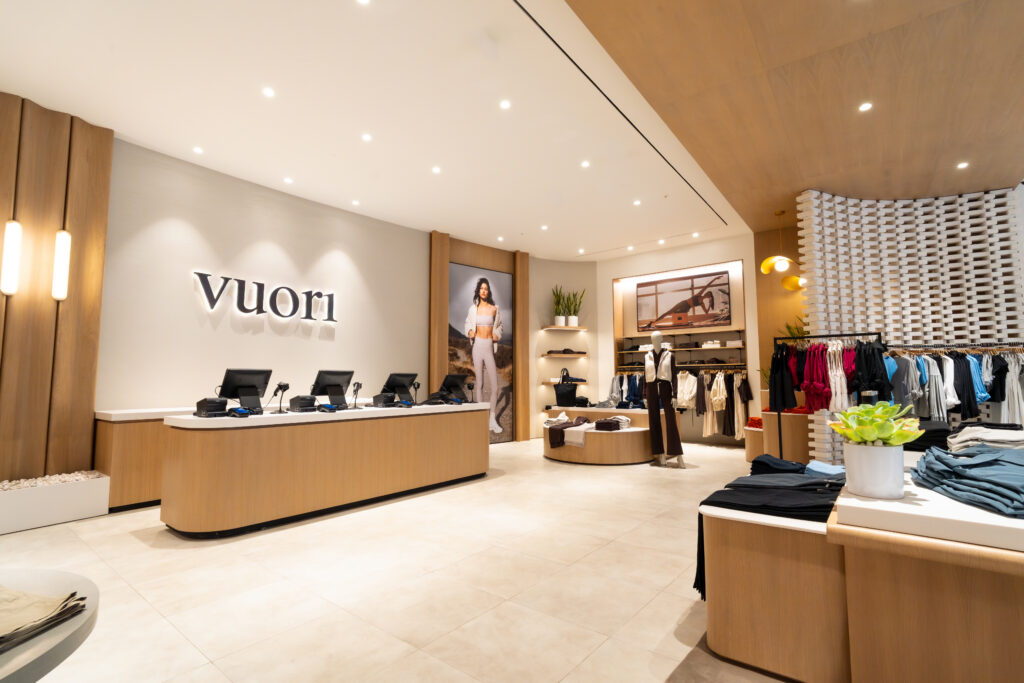 VUORI-D2-STORE-2 - Petróleo y Energía Bright clothing store interior with a curved wood checkout counter, Vuori sign on the wall, and racks of apparel on display.