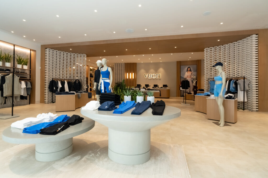 VUORI-D2-STORE-4 - Petróleo y Energía Spacious clothing store interior with circular display tables of folded blue and black apparel and mannequins wearing blue athletic outfits.