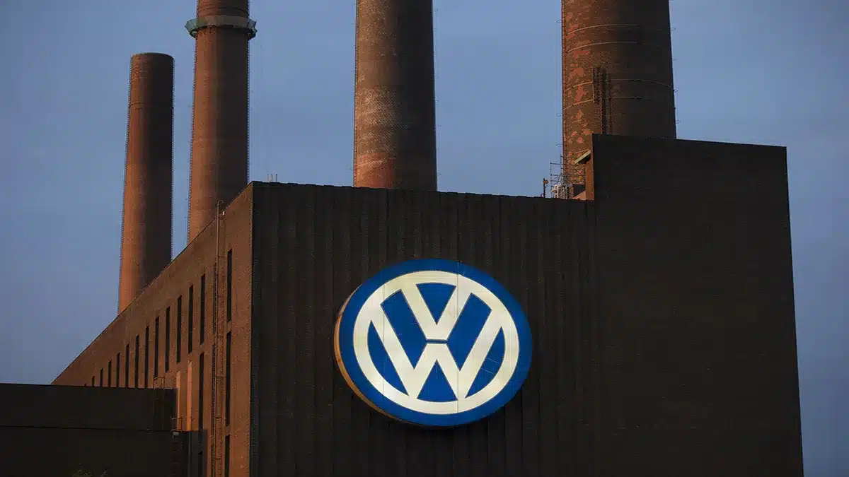 Volkswagen factory building with a large VW circular logo on a dark wall and tall smokestacks in the background.