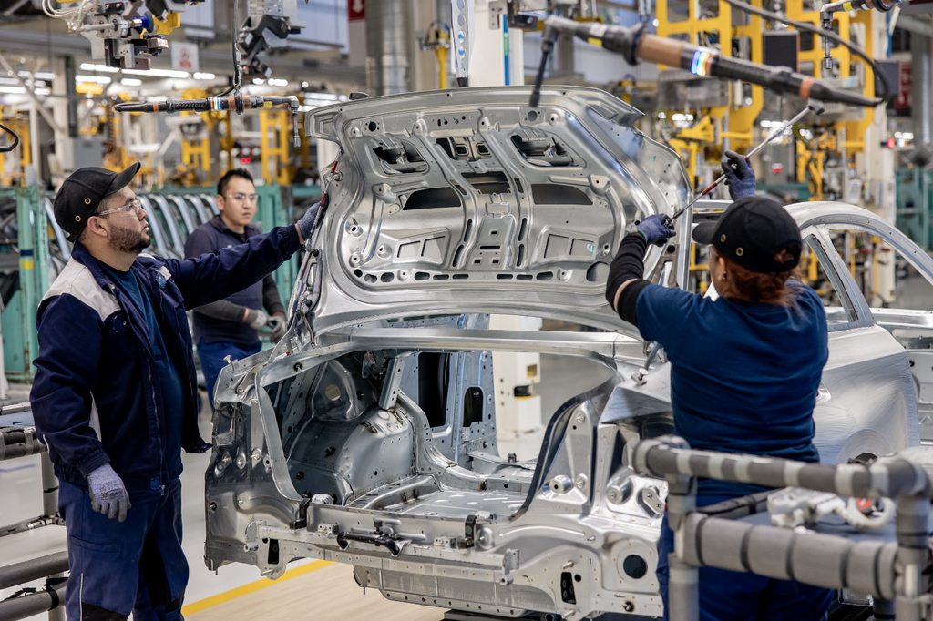 WhatsApp Image 2026-09-08 at 94525 AM 1 - Petróleo y Energía Three factory workers in blue uniforms install a silver car body shell on an automotive assembly line.