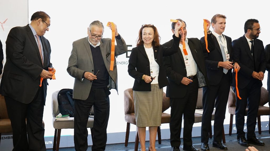 WhatsApp Image 2026-09-11 at 82528 AM - Petróleo y Energía Group of professionals on a stage holding orange ribbons up, mid ribbon-cutting ceremony at a conference.