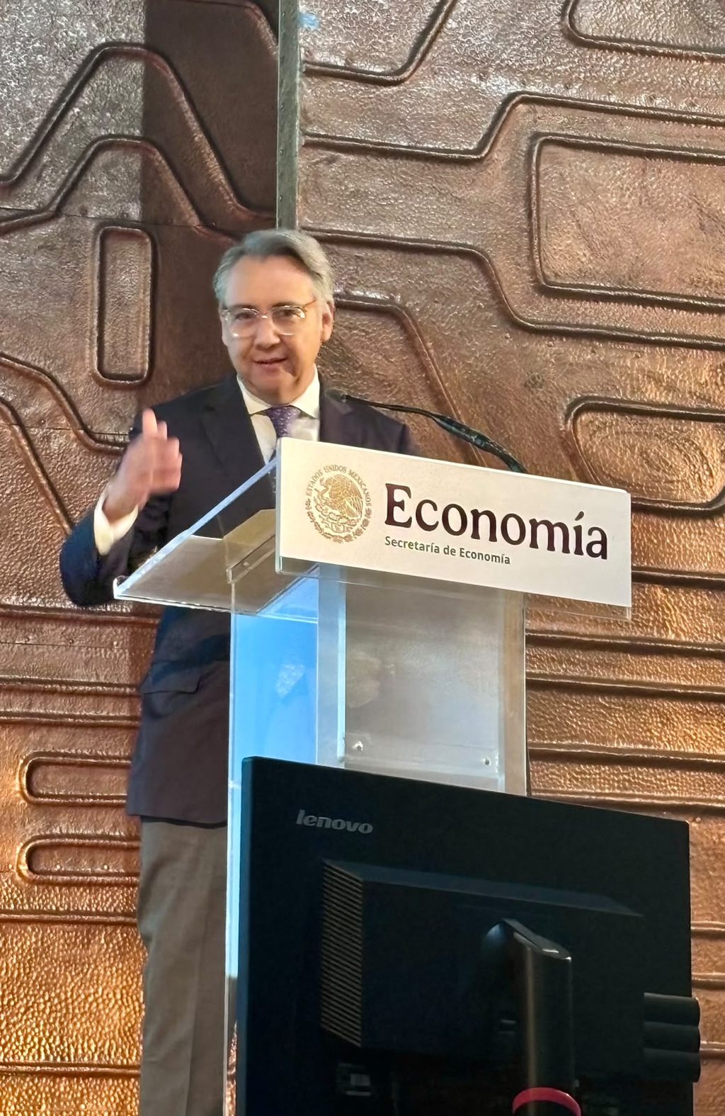 WhatsApp Image 2026-09-15 at 094534 - Petróleo y Energía Man in a dark suit giving a speech at a podium labeled Economía, Secretaría de Economía, with a textured bronze wall behind him.