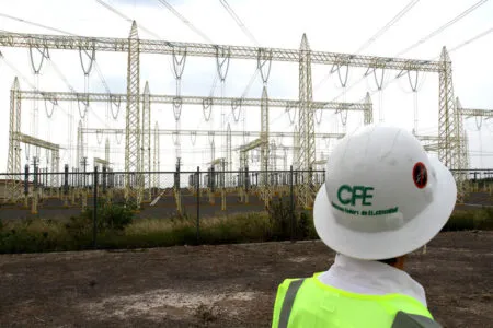 Worker in a high-visibility vest standing before an electrical substation with yellow lattice towers and power lines; helmet reads 'CFE' (Mexico's electric utility).