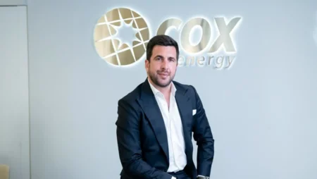 Man in a navy suit sits in front of a COX Energy logo on a light-gray wall in an office setting.