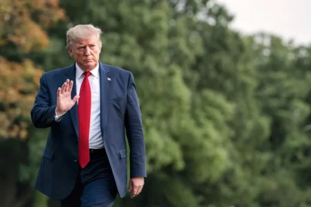 Man in a navy suit with a red tie walks outdoors, raising his right hand.