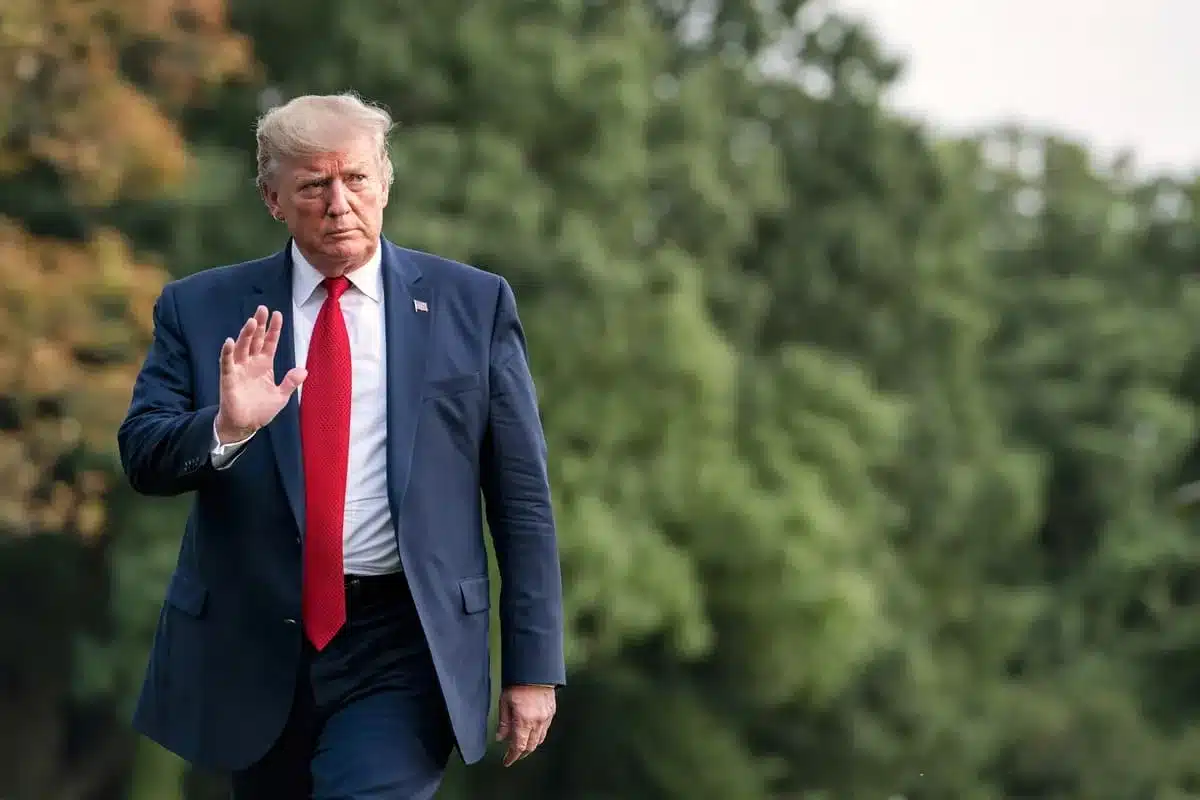 Man in a navy suit with a red tie walks outdoors, raising his right hand.