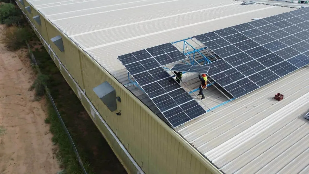 Aerial view of workers installing a large solar panel array on a beige industrial roof with a scaffold platform nearby