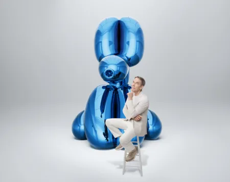 Man in a light-colored suit sits on a small ladder next to a tall glossy blue abstract sculpture made of balloon-like shapes.
