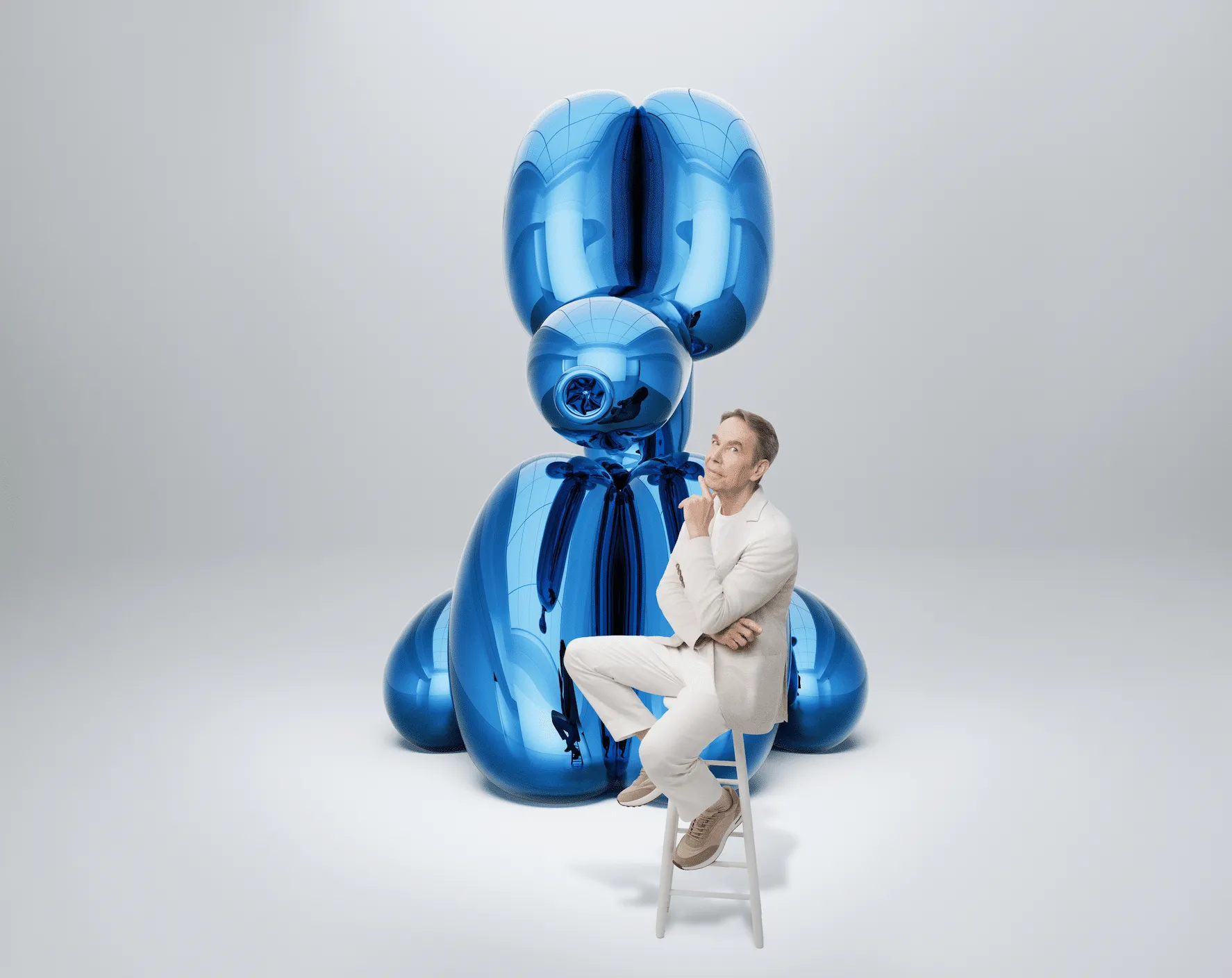 Man in a light-colored suit sits on a small ladder next to a tall glossy blue abstract sculpture made of balloon-like shapes.