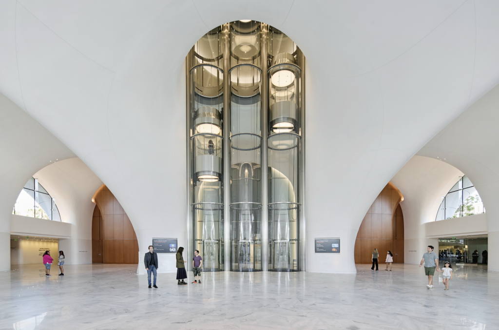 museo lucas 2026-09-23 a las 94806 am - Petróleo y Energía Museum or public atrium with a tall glass elevator shaft between curved white walls and a marble floor, several people nearby for scale.