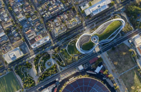 Aerial view of a dense urban area featuring a futuristic white looping building with green roofs and surrounding roads and parks, including a large circular stadium at the bottom right.