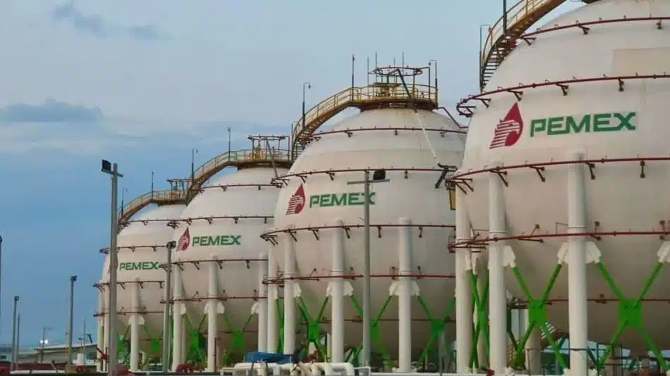 Row of large white PEMEX storage tanks with green supports at an industrial facility, pipelines and scaffolding visible in the foreground.