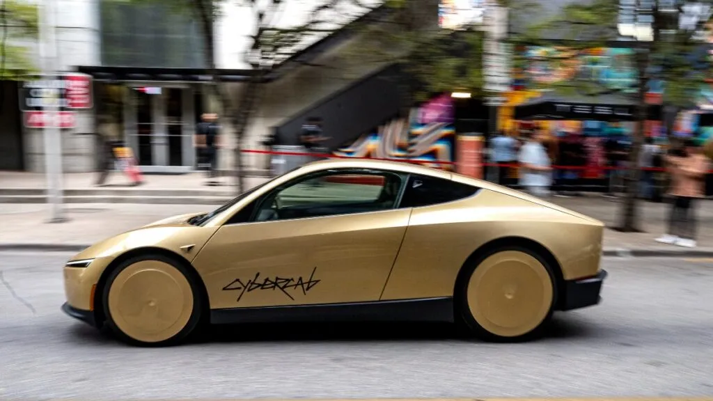 Gold-colored sports car driving quickly down a city street with blurred pedestrians and storefronts in the background.