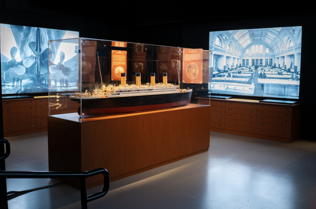 titanic 2026-09-28 a las 93823 am - Petróleo y Energía Scale model of a large ocean liner displayed in a glass case on a wooden stand, with blue-toned panels showing shipyard and interior scenes behind it