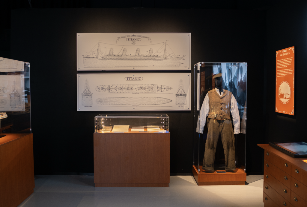 titanic 2026-09-28 a las 93838 am - Petróleo y Energía Two large Titanic blueprints on a dark wall, with a glass case containing a period suit in front and an orange information panel to the right.