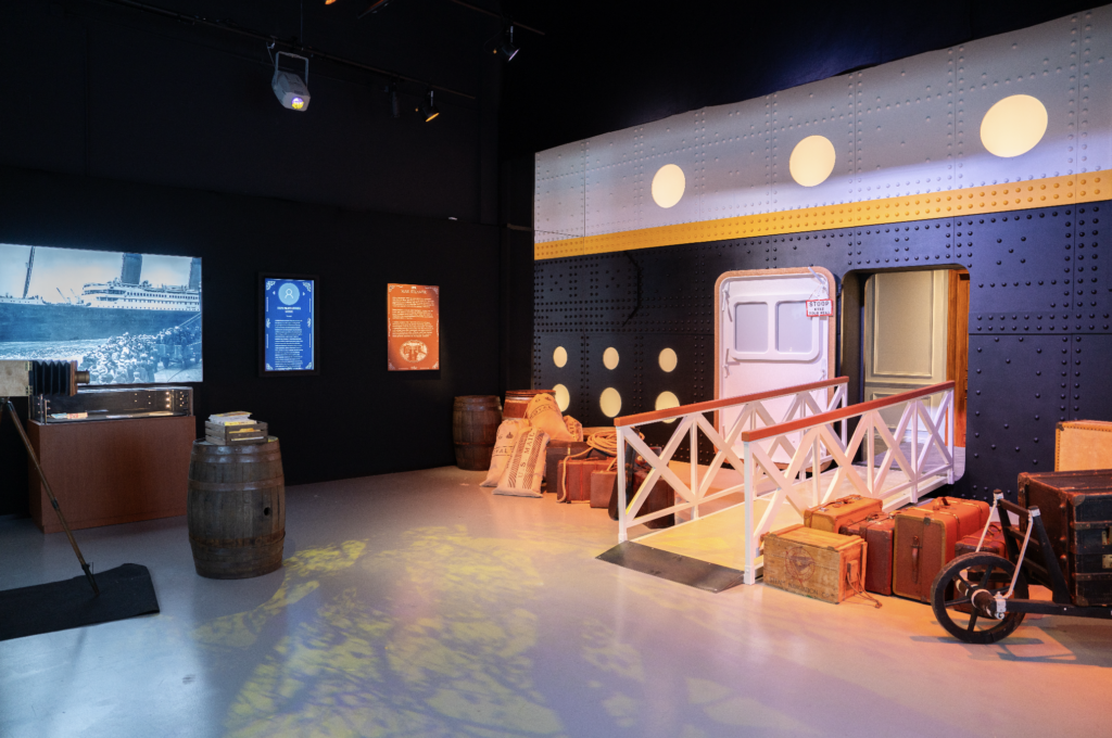 - Petróleo y Energía Museum exhibit of a ship deck scene with a blue riveted wall, yellow stripe, circular lights, and a white railing, with crates, barrels and sacks in the foreground.