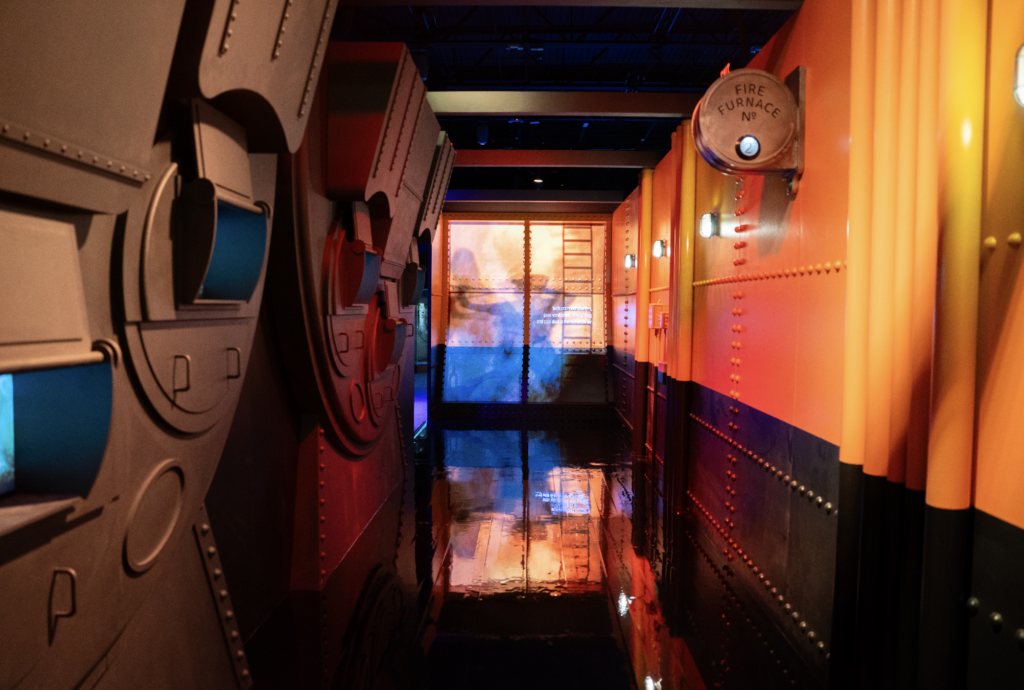 titanic 2026-09-28 a las 94002 am - Petróleo y Energía Industrial-looking exhibit hallway with riveted metal walls in gray on the left and orange-yellow panels on the right; a circular'Fire Furnace No. 2' gauge mounted on the right wall.