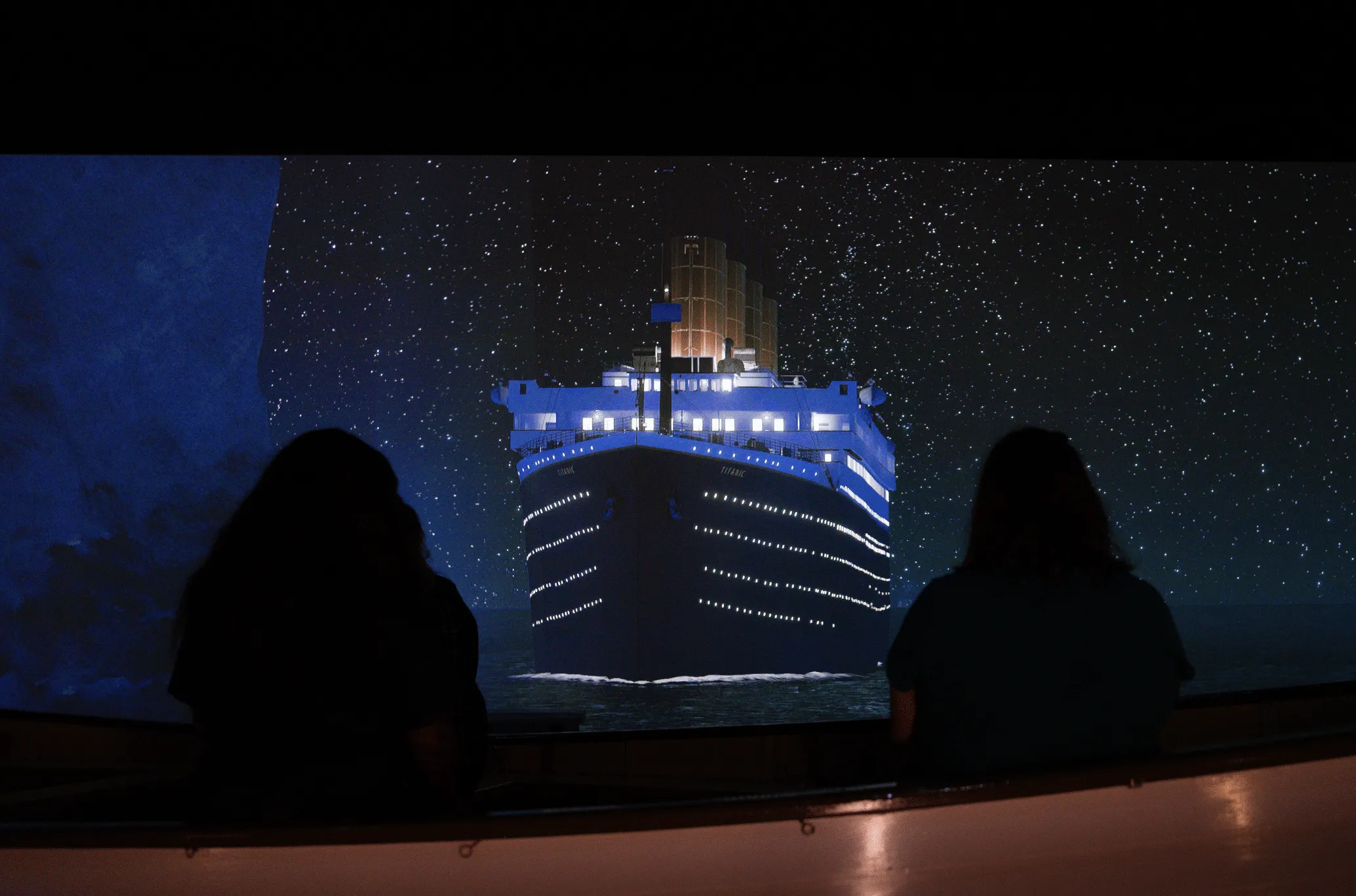 Large cruise ship model illuminated against a starry backdrop, viewed from a foreground audience silhouette.