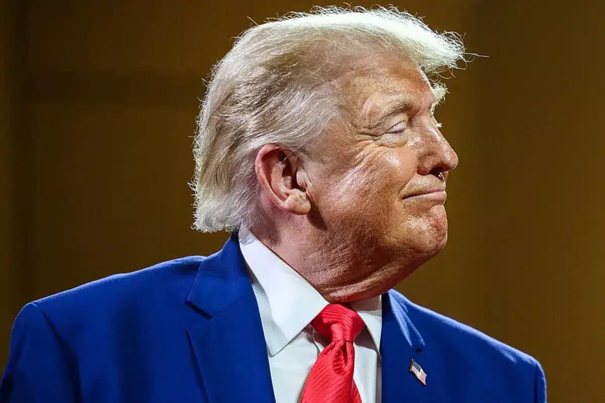 Smiling man in a blue suit with a red tie, seen in profile to the right, wearing an American flag lapel pin.