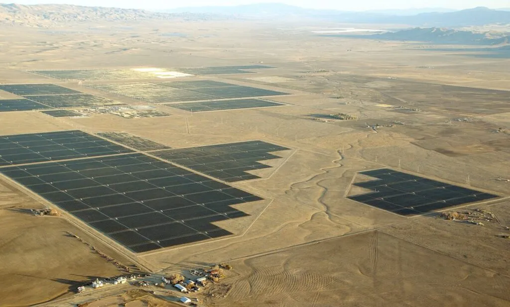A vast desert solar farm with numerous dark photovoltaic panels arranged in rectangular fields.
