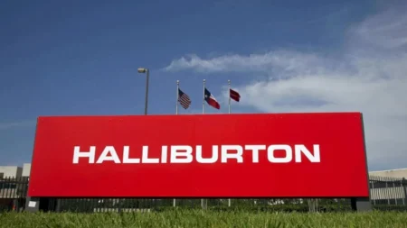 Red Halliburton company sign with white lettering, set on a grassy foreground and flagpoles with US and Texas flags against a blue sky.