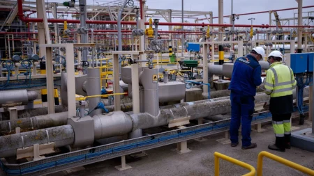 Two workers in hard hats and high-visibility vests inspect a complex network of pipes and valves in an industrial plant.