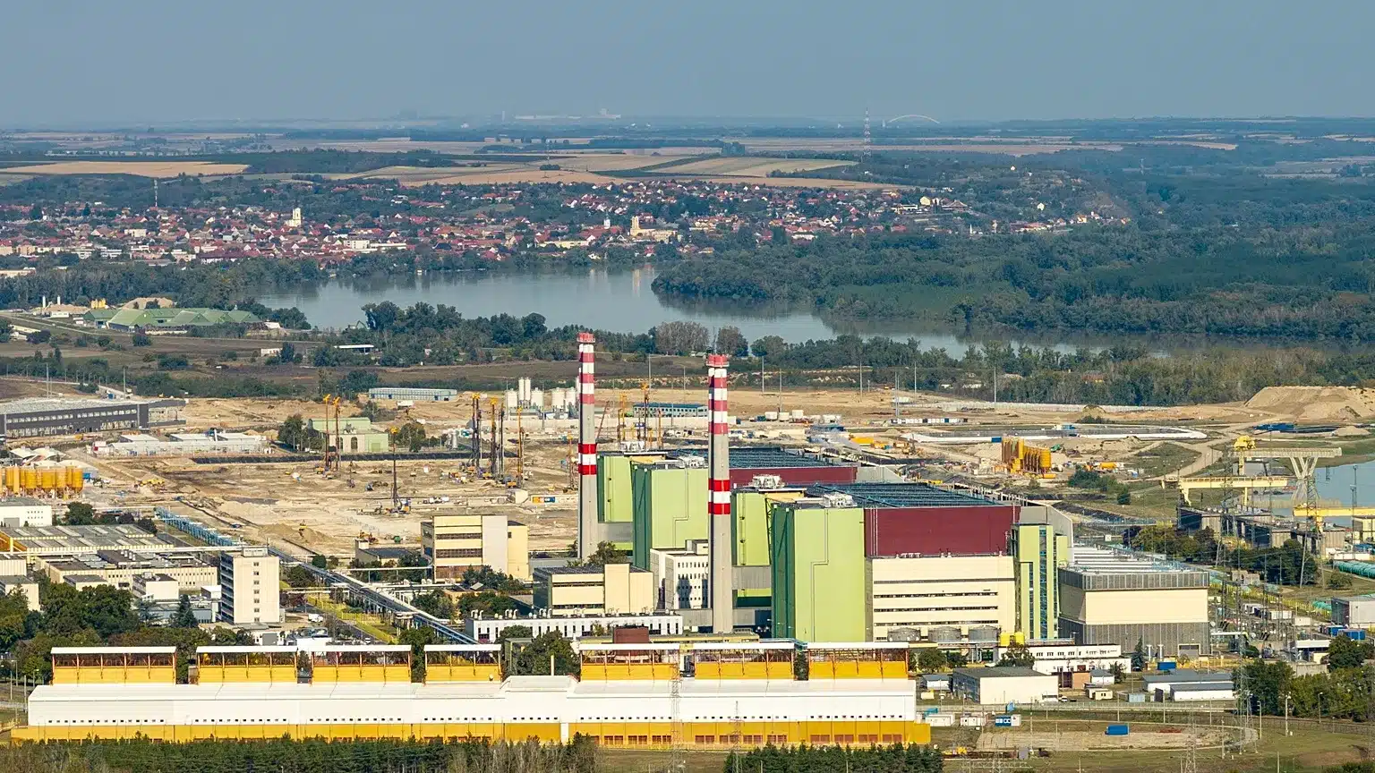 Industrial facility with tall striped smokestacks and green-and-beige buildings, set near a river and distant town.