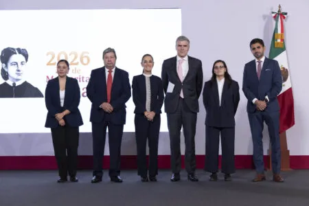 Six professionally dressed individuals stand in a row on a stage for a formal group photo, with a large white screen on the left and a Mexican flag on the right behind them.