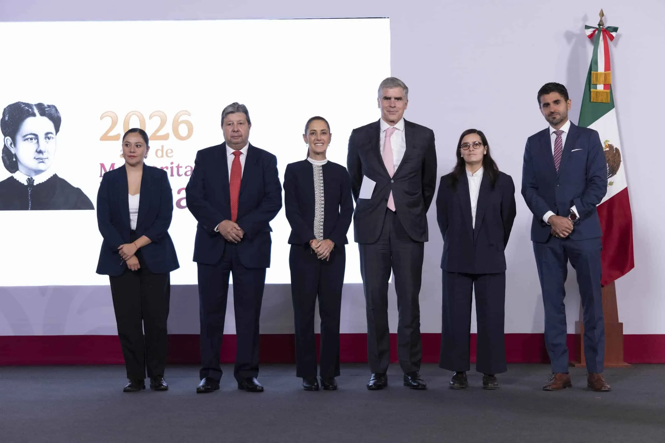 Six professionally dressed individuals stand in a row on a stage for a formal group photo, with a large white screen on the left and a Mexican flag on the right behind them.