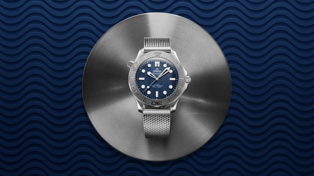 21030422003004_BondSpectre_ADV_SE_KV_NoLogo_16_9_RVB - Petróleo y Energía Omega Seamaster stainless steel watch with a blue dial on a brushed metal disc, set against a blue wavy background.