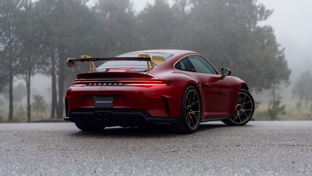 25 - Petróleo y Energía Rear three-quarter view of a red Porsche 911 GT3 with a large rear wing and gold-rimmed wheels, parked on a wet road with a misty, forested background.