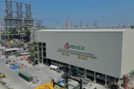 Industrial processing facility with a large gray building labeled PEMEX, diesel hydrodesulfurization plant under construction, cranes and equipment nearby