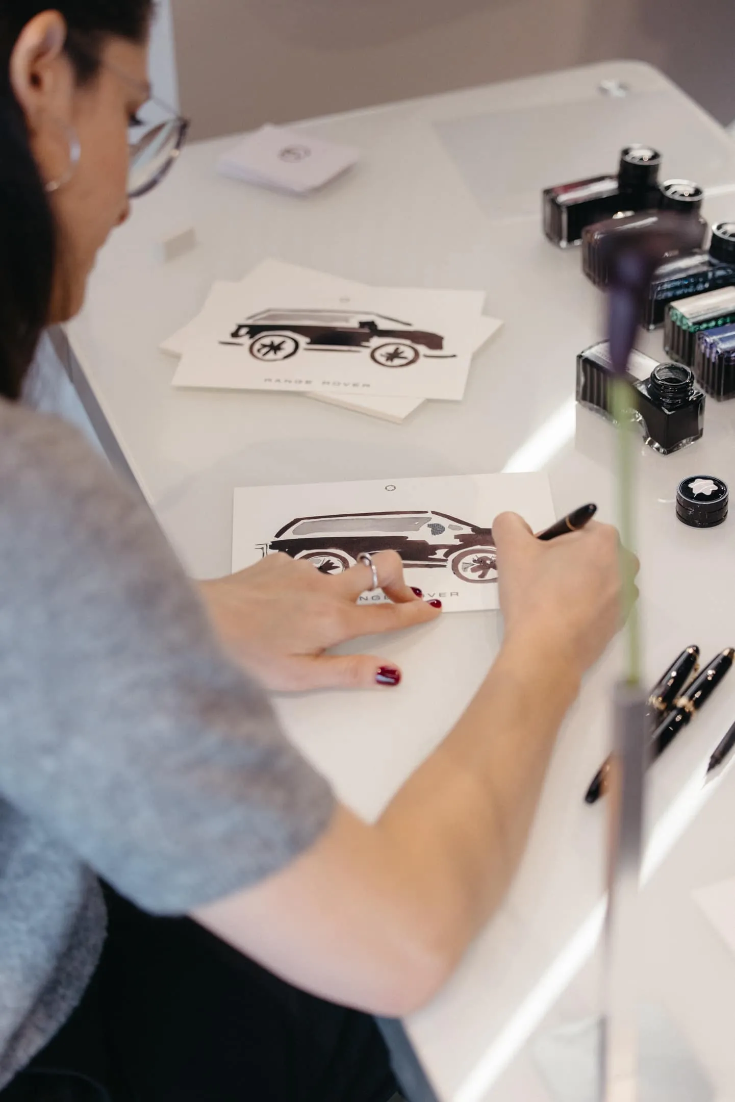 Person sketching a vintage car design on white card at a studio desk, with ink bottles and drawing tools nearby.