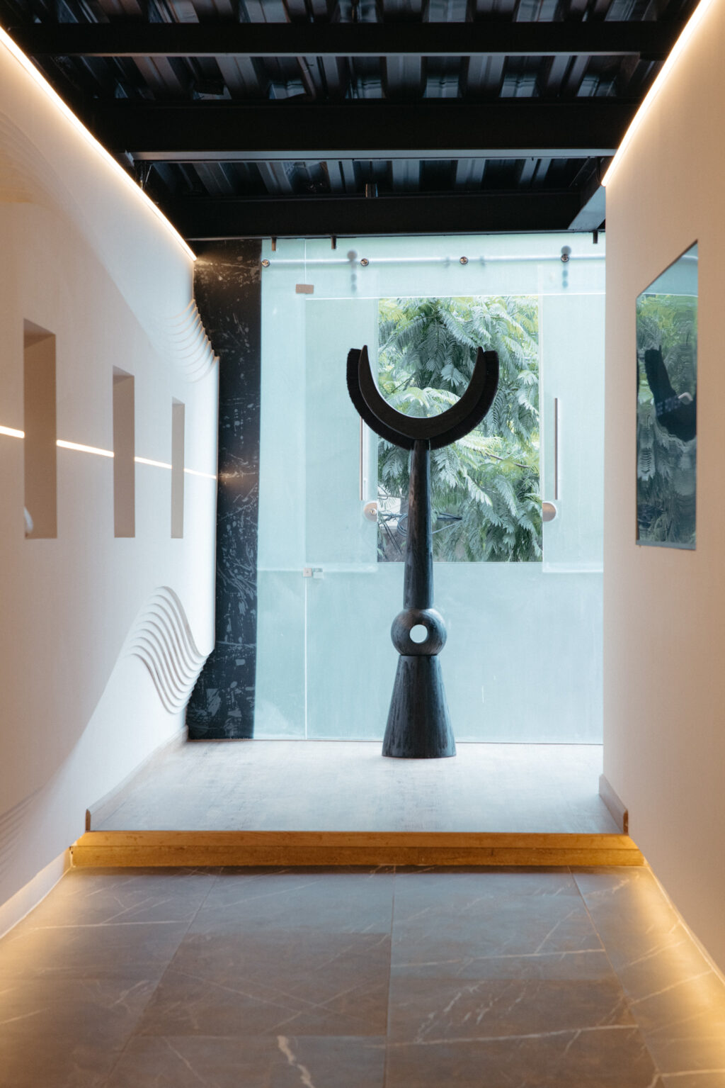 Range Rover - Petróleo y Energía Interior hallway with a tall black curved sculpture on a pedestal, glass wall and greenery outside visible beyond.