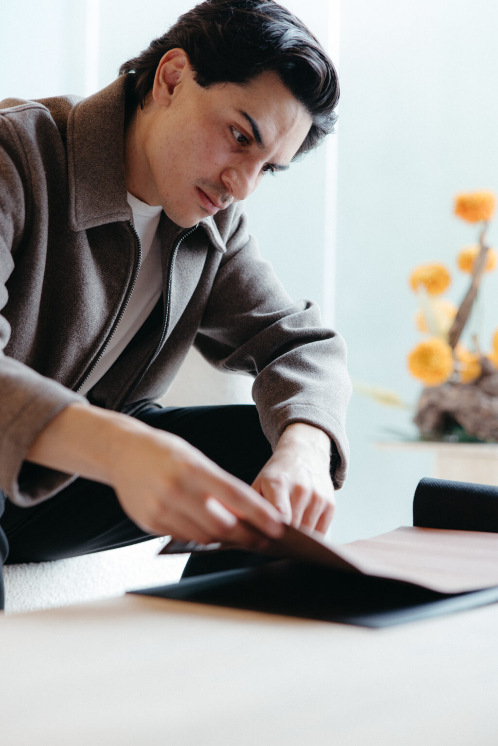 - Petróleo y Energía Man in a brown jacket leaning over a table, writing on a document with a focused expression in a bright room.