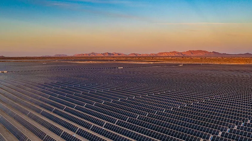 Rows of solar panels stretching across a desert plain at sunset with distant mountains on the horizon, pastel sky above.