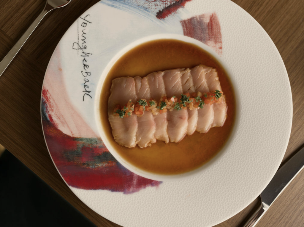 5 EXCUSAS GOURMET 2026-10-15 a las 32233 pm - Petróleo y Energía Sliced tuna tataki arranged in a shallow orange sauce in a white bowl on a decorative plate, with a knife nearby on a wooden table.