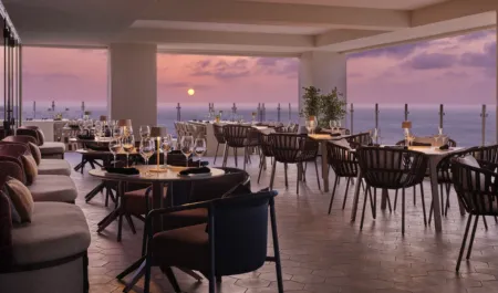 Elegant seaside rooftop restaurant at sunset with tables set for dinner and an ocean view beyond glass railing.
