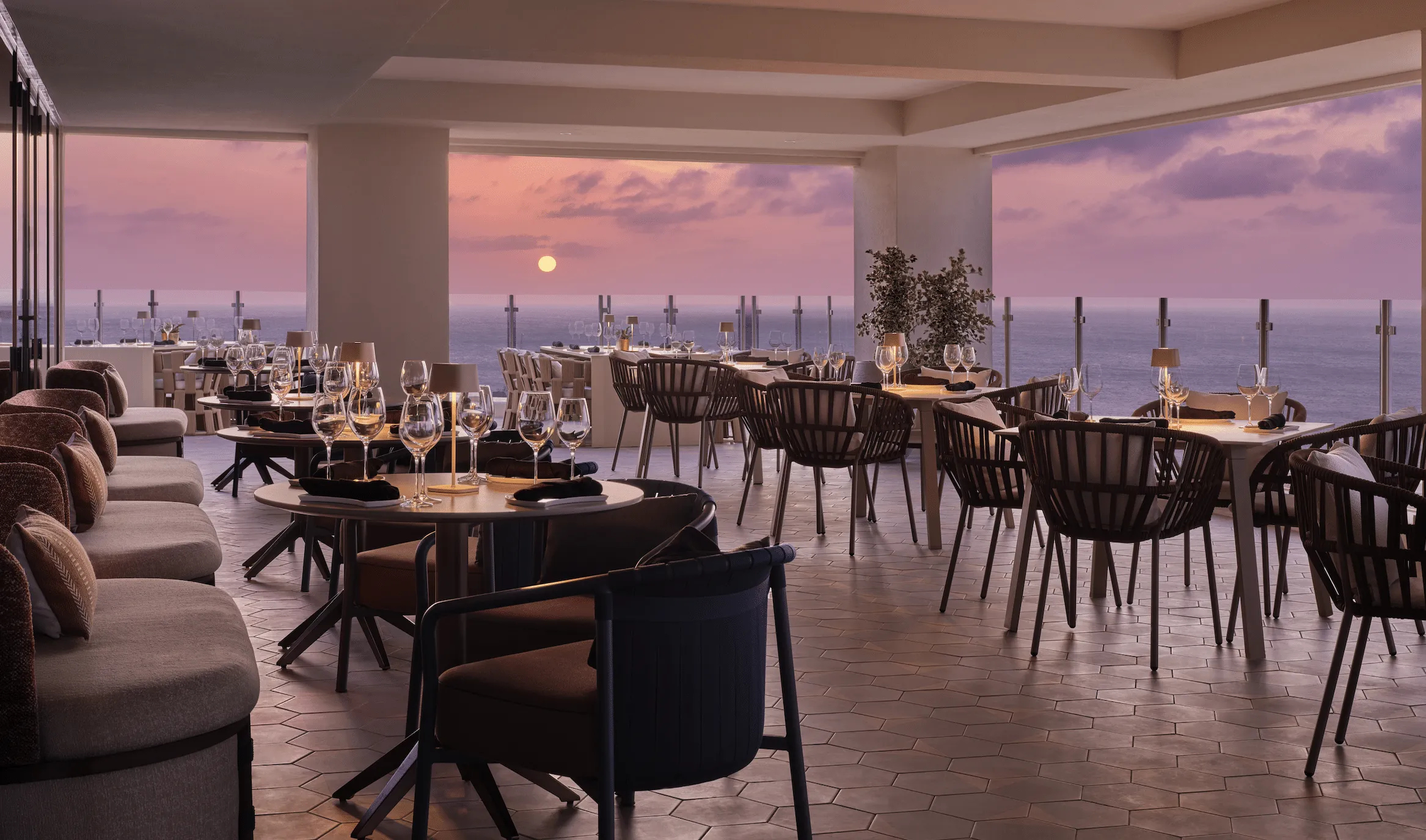 Elegant seaside rooftop restaurant at sunset with tables set for dinner and an ocean view beyond glass railing.