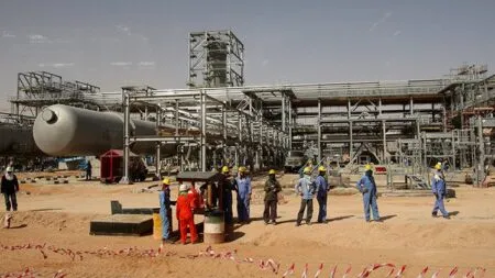 Construction workers in blue and orange coveralls with hard hats stand near large industrial pipelines at an oil refinery under construction.