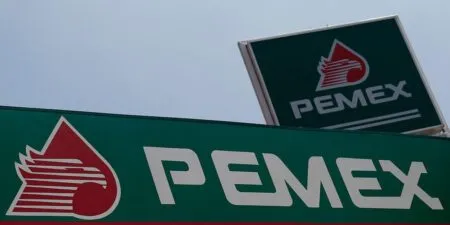 Pemex logo on a green sign with a red droplet graphic and bold white letters 'PEMEX' at a tilted angle in the foreground and a smaller sign in the background.