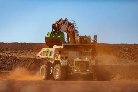 Large mining haul truck on a dusty site with an excavator attachment on its upper deck, kicking up orange dust in a barren landscape.