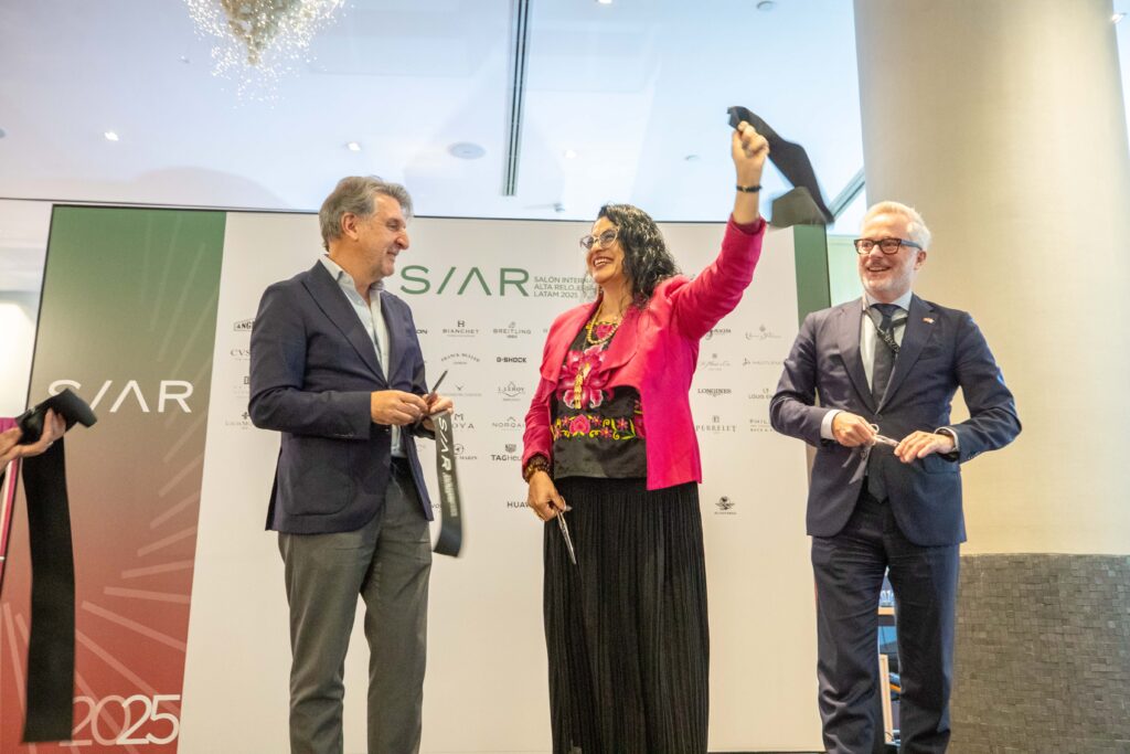 A10I0215 1 - Petróleo y Energía Three people on a stage at an award event; the woman in a bright pink blazer raises a dark trophy with a smile, flanked by two men in suits.